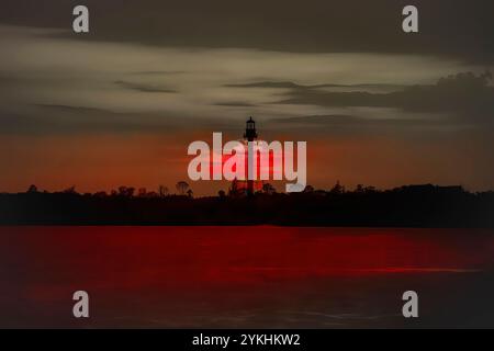 Dramatic red sunset behind a silhouetted lighthouse with reflection across calm water Stock Photo