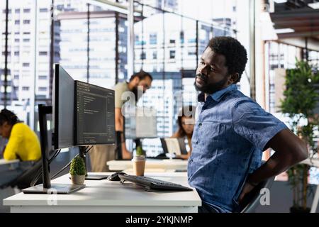 Admin stretching to relieve muscle strain, conducting tests to optimize machine learning models for performance and accuracy. IT expert in office suffering back pain from bad posture, analyzing data Stock Photo