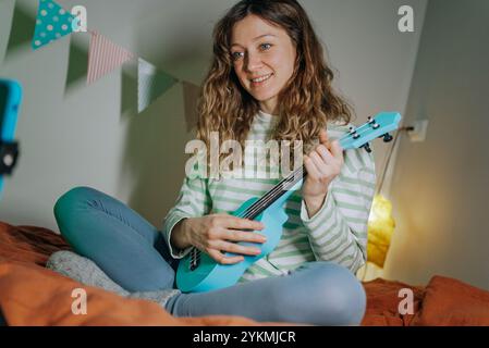Young woman musician happily plays the ukulele guitar, recording a video lesson or song cover with her smartphone at home Stock Photo