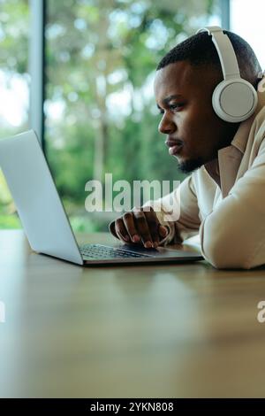 A focused young African man wearing headphones is working on his laptop. He is deeply engaged with the task at hand, conveying a sense of concentratio Stock Photo