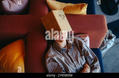 A tired student lies on a sofa with a book on their face, illustrating exhaustion from studying hard in a comfortable home setting. Stock Photo