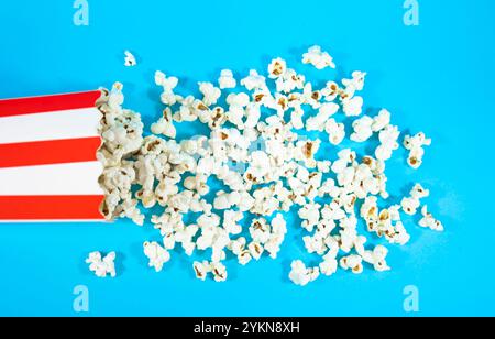 Top view of fallen red and white cardboard bucket scattering popcorn on a blue background, cinema food concept Stock Photo