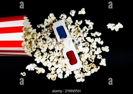 Top view of red and white cardboard bucket lying with 3D glasses on top of scattered popcorn on black background, popcorn and cinema concept Stock Photo