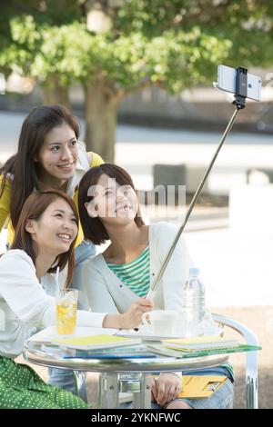 Three female university students taking selfie stick photos in campus cafeteria Stock Photo