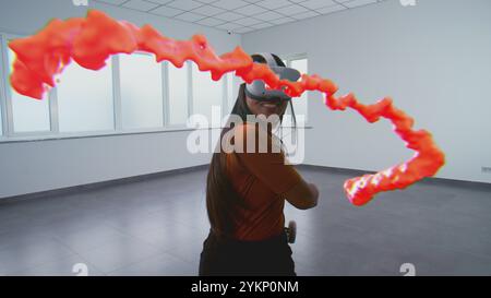 Creative Asian female artist in augmented reality headset paints with wireless controllers, uses gestures to create abstract online concept art. Holographic 3D graphics of paint drops. VFX animation. Stock Photo