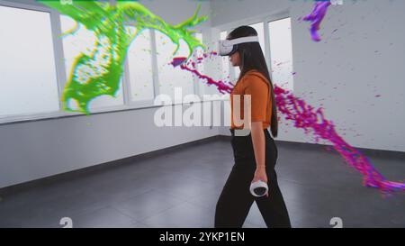 Asian young woman in virtual reality headset paints with wireless controllers, uses gestures to create abstract online artwork. Holographic 3D animation of paint drops. Augmented reality technology. Stock Photo