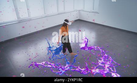 Talented Asian young woman wearing VR headset paints with wireless controllers, uses gestures to create abstract online artwork. Holographic 3D rendering of paint drops. Augmented reality technology. Stock Photo