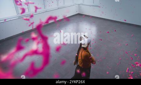 Talented Asian young woman wearing VR headset paints with wireless controllers, uses gestures to create abstract online artwork. Holographic 3D rendering of paint drops. Augmented reality technology. Stock Photo