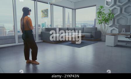 Talented Asian female designer in augmented reality headset uses virtual interior design software to arrange stylish apartment or office room with 3D furniture and equipment. Futuristic VFX animation. Stock Photo