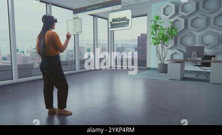 Asian female designer wearing virtual reality headset uses AI augmented reality interior design app to arrange office workspace or stylish apartment with 3D furniture and equipment. 3D visual effects. Stock Photo
