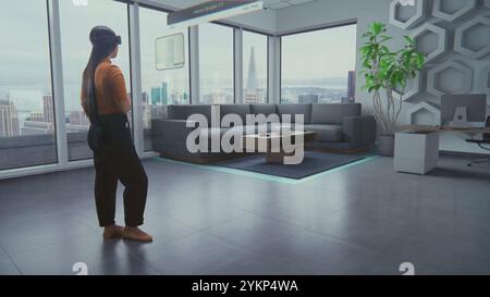 Asian female designer wearing virtual reality headset uses AI augmented reality interior design app to arrange office workspace or stylish apartment with 3D furniture and equipment. 3D visual effects. Stock Photo
