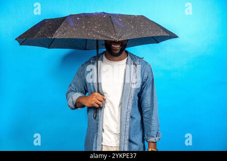 Stylish young man is holding an umbrella covering his face and smiling on a blue background Stock Photo