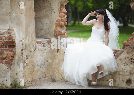 The bride in an elegant white dress gracefully poses against the stunning picturesque vintage background of the ruined fortress wall Stock Photo
