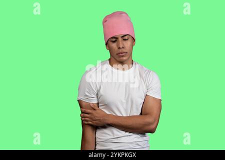 Pensive young latino man with pink beanie and white t-shirt on chroma key background, holding his arm and looking down Stock Photo