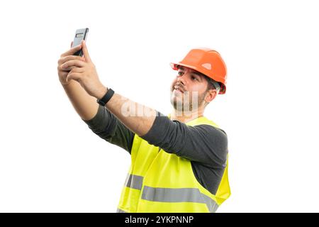 Friendly constructor wearing fluorescent work vest and orange safety hardhat taking selfie using smartphone camera isolated on white studio background Stock Photo