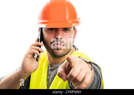 Trustworthy builder man wearing hardhat and work vest uniform talking on phone pointing index finger at camera isolated on white studio background Stock Photo