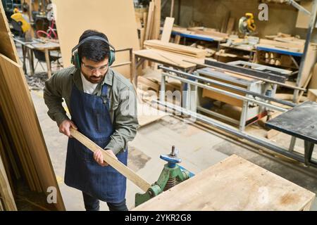 A focused young carpenter operates machinery in a bustling lumberyard. Wearing safety gear, he embodies precision and dedication, contributing to team Stock Photo