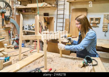 A young woman wears safety goggles while using a drill to assemble a wooden chair in a lumberyard workshop. The scene highlights craftsmanship, woodwo Stock Photo