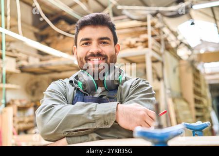 A young man stands confidently in a busy lumberyard, wearing headphones and work attire. His smile reflects the spirit of teamwork and skill developme Stock Photo