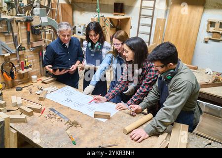 A diverse group of young apprentices collaborates with an experienced mentor in a busy woodworking shop. They focus intently on project plans, demonst Stock Photo