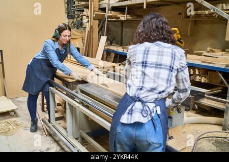 Two young women work together in a lumberyard, showcasing teamwork and skill. They're actively engaged in woodworking, wearing protective gear amidst Stock Photo
