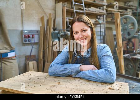 A young woman wearing a denim shirt smiles confidently in a workshop surrounded by wood and machinery, embodying the spirit of craftsmanship and indus Stock Photo