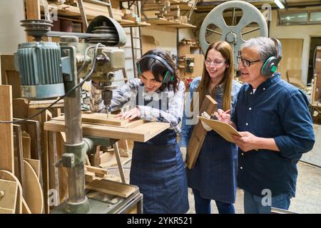 Experienced mentor guiding apprentices in a busy lumberyard. The team demonstrates woodworking skills, safety, and collaboration in a workshop setting Stock Photo
