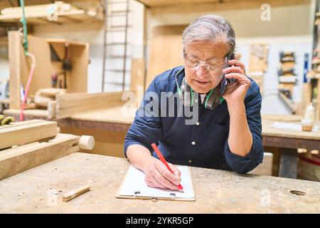 An older carpenter wearing safety glasses and headphones is multitasking in a woodworking shop, talking on the phone and writing on a clipboard. Stock Photo