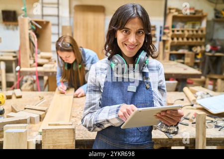 A smiling young woman using a tablet in a bustling lumberyard, wearing headphones and apron. Another worker focuses on a wooden plank nearby, showcasi Stock Photo