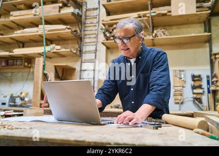 An experienced craftsman is focused on using a laptop in a well-organized lumber workshop, surrounded by woodworking tools and materials. The image ca Stock Photo