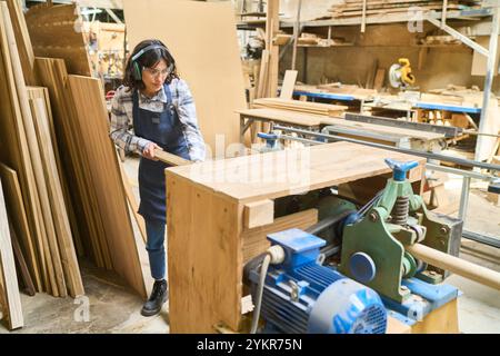 A young woman operates woodworking machinery in a busy lumberyard. The scene depicts skilled craftsmanship and apprenticeship in a dynamic setting, fo Stock Photo