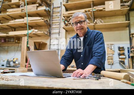 Mature craftsman in a wood workshop using a laptop on a workbench surrounded by tools and wooden materials, showcasing a blend of traditional craftsma Stock Photo