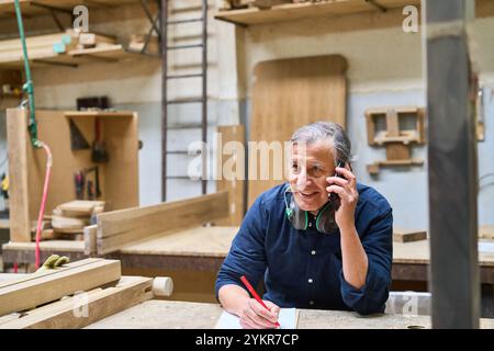 An experienced lumberyard worker enthusiastically discusses work on the phone while taking notes. The scene captures professionalism, communication, a Stock Photo
