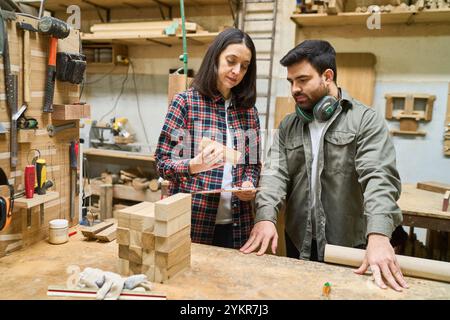 Two professionals engage in discussion while working with wood materials. Focus on teamwork and training in a busy lumberyard environment, showcasing Stock Photo