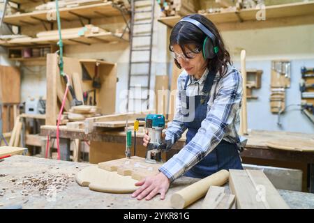 A focused worker using a router on a wooden piece in a bustling woodworking shop. Safety equipment and organized workspace highlight professionalism a Stock Photo