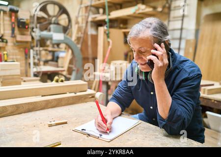 An older craftsman is making notes while speaking on the phone in a woodworking workshop, surrounded by tools and wood. The scene captures the industr Stock Photo