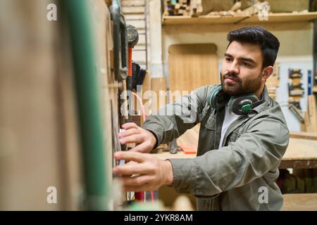 A young man wearing headphones operates machinery in a lumberyard. He is focused and wearing a work jacket, highlighting a scene of industrial work an Stock Photo