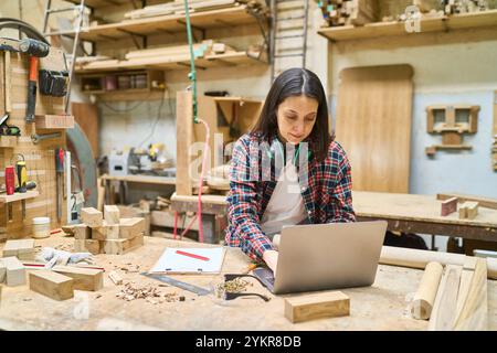A young woman concentrates on her laptop amid a bustling lumberyard, showcasing teamwork and craftsmanship. Tools and wooden materials surround her, i Stock Photo
