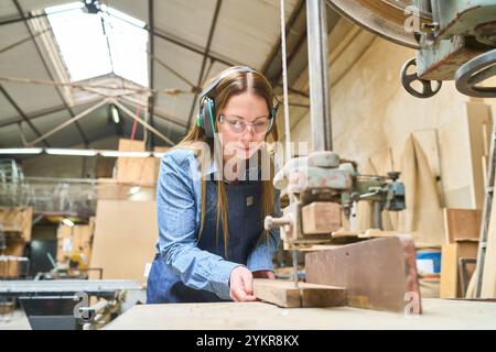 A young woman focuses on operating machinery in a lumberyard, embodying themes of concentration, craftsmanship, and industrial work. She wears safety Stock Photo