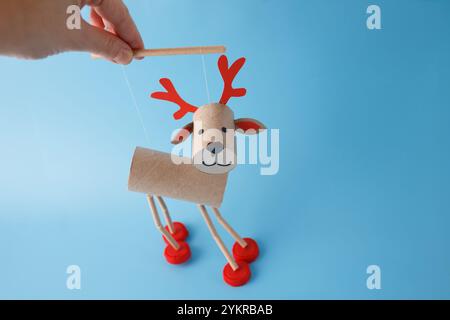 Hand holding DIY reindeer puppet made of cardboard tubes, plastic caps, and paper. on a blue background. Christmas craft and holiday decoration concep Stock Photo