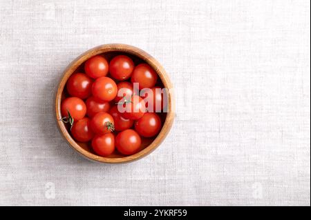 Small red cherry tomatoes in a wooden bowl on linen fabric, from above, with empty space for text. Fresh, ripe, tiny and round cocktail tomatoes. Stock Photo