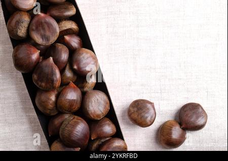 Sweet chestnuts in cardboard punnet on linen, from above, with empty space for text. Castanea sativa, raw, whole and unshelled. Stock Photo