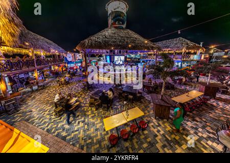 The Pasawá Box Eatery on Caracasbaaiweg in Willemstad, Curaçao, is seen at night: the dining area, built from shipping containers, combines thatched pavilions, a central bar and a colourfully paved outdoor area. The wall decoration is by Sander van Beusekom (artist name BLEND); in the background, a painted cylindrical container serves as a room divider. Stock Photo