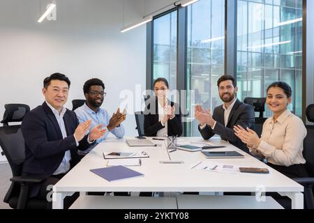 Positive business environment in office. Diverse group of professionals in meeting room clapping, signifying teamwork, success, and collaboration. Phones and documents on table. Stock Photo