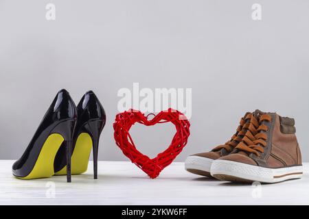 Red knitted heart with black womens high heel shoes and mens sneakers. Footwear on white background with copy space Stock Photo