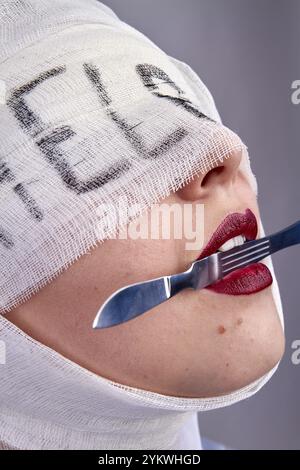 Close up womans face holding scalpel in her teeth. Bandaged head with help word Stock Photo