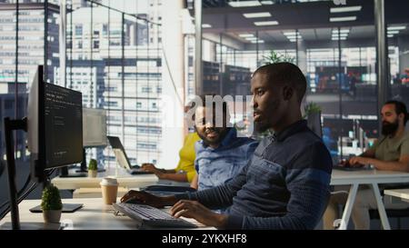 Programmer doing software debugging, asking for help from smiling team leader in wheelchair. Employee fixing coding mistakes, being assisted by cheerful manager with disability, camera B Stock Photo