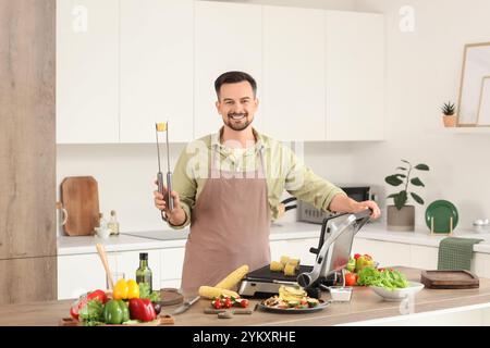 Young happy man cooking fresh corn cobs on modern electric grill in kitchen Stock Photo