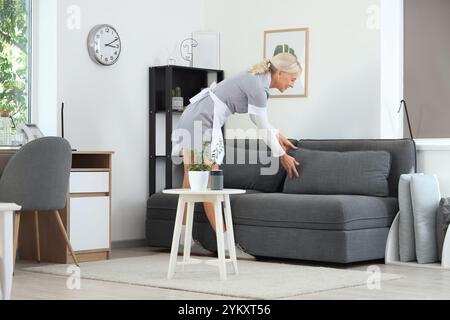 Middle-aged chambermaid adjusting pillows on sofa in hotel room Stock Photo