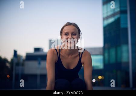 Young woman smiling while practicing yoga outdoors at sunset near modern buildings Stock Photo